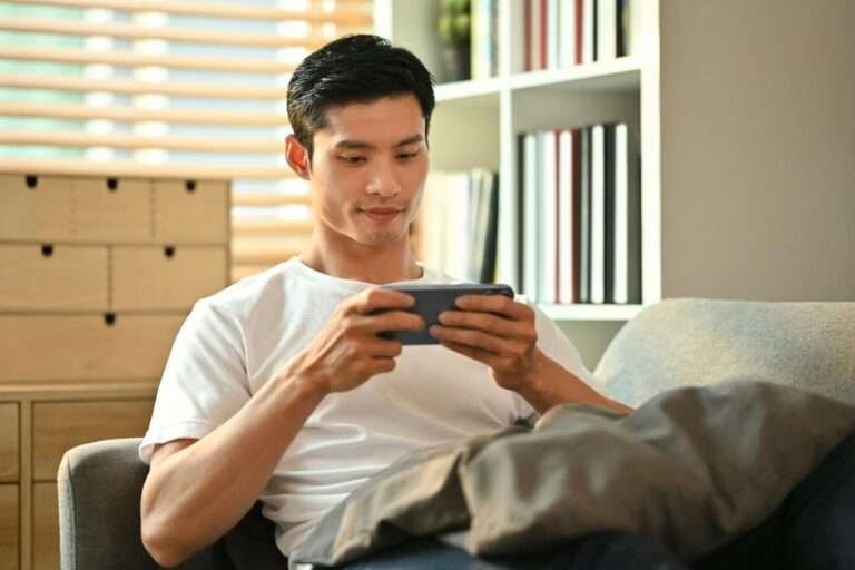 Happy millennial man playing video games on smart phone while resting on couch.